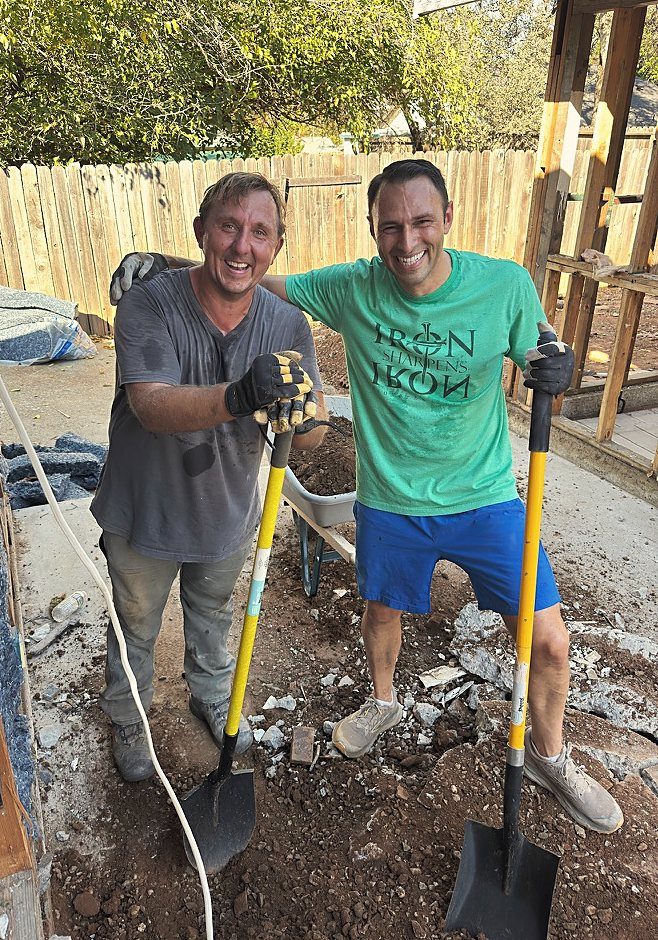 Daniel and Russ smiling with shovels on a renovation site, Russ wearing an Iron Sharpens Iron shirt